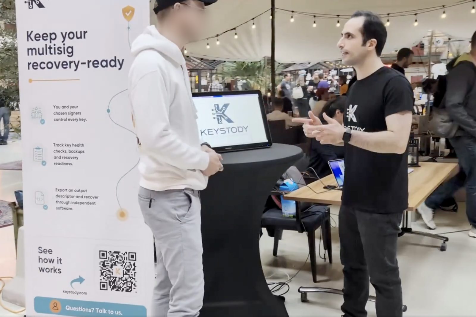 A conversation at the Keystody booth: two people talking beside the roll-up banner and a screen showing the Keystody logo